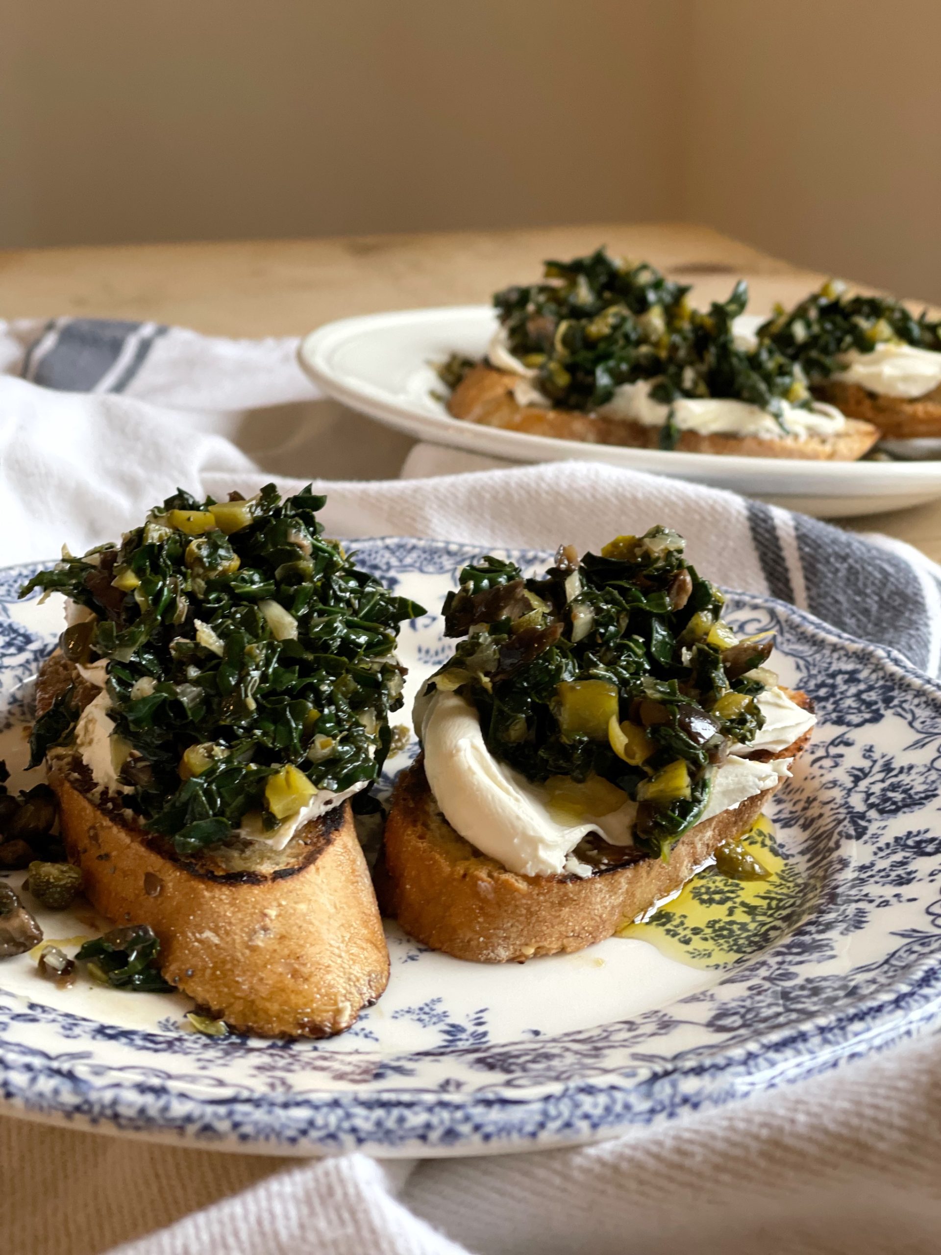 A recipe for kale bruschetta. A mixture of kale or cavelo nero with capers, olives, cornichons and herbs on a garlicky toasts with soft goat's cheese